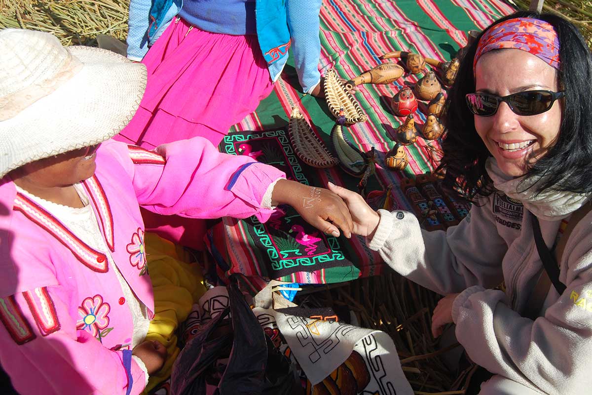Dos mujeres, una de ellas con gafas de sol, se dan la mano ante un colorido expositor textil con artesanía hecha a mano en un mercado al aire libre.