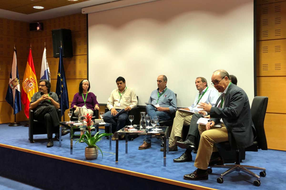 Siete personas sentadas en un panel del escenario frente a una pantalla de proyección, con banderas y micrófonos visibles; parecen estar participando en un debate o una presentación.