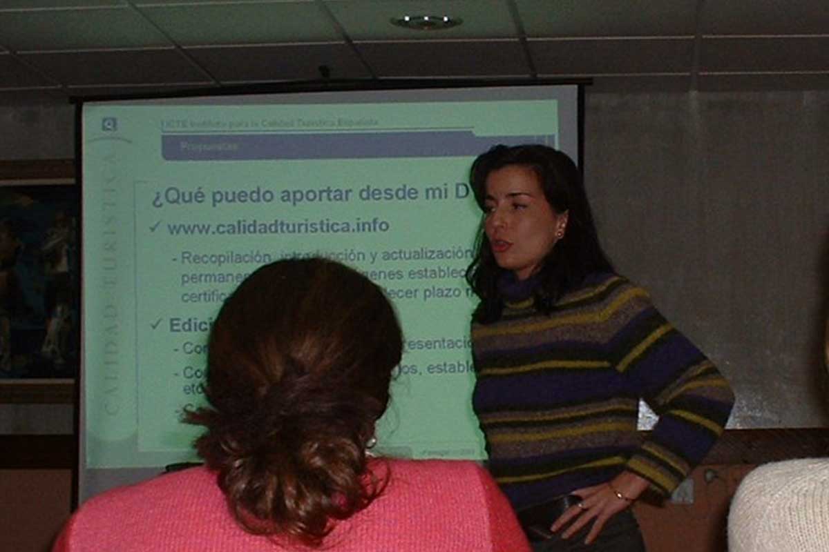 Una mujer habla de pie delante de una diapositiva proyectada en una sala de conferencias. El texto de la diapositiva está en español y habla de las contribuciones y actualizaciones en materia de calidad turística.