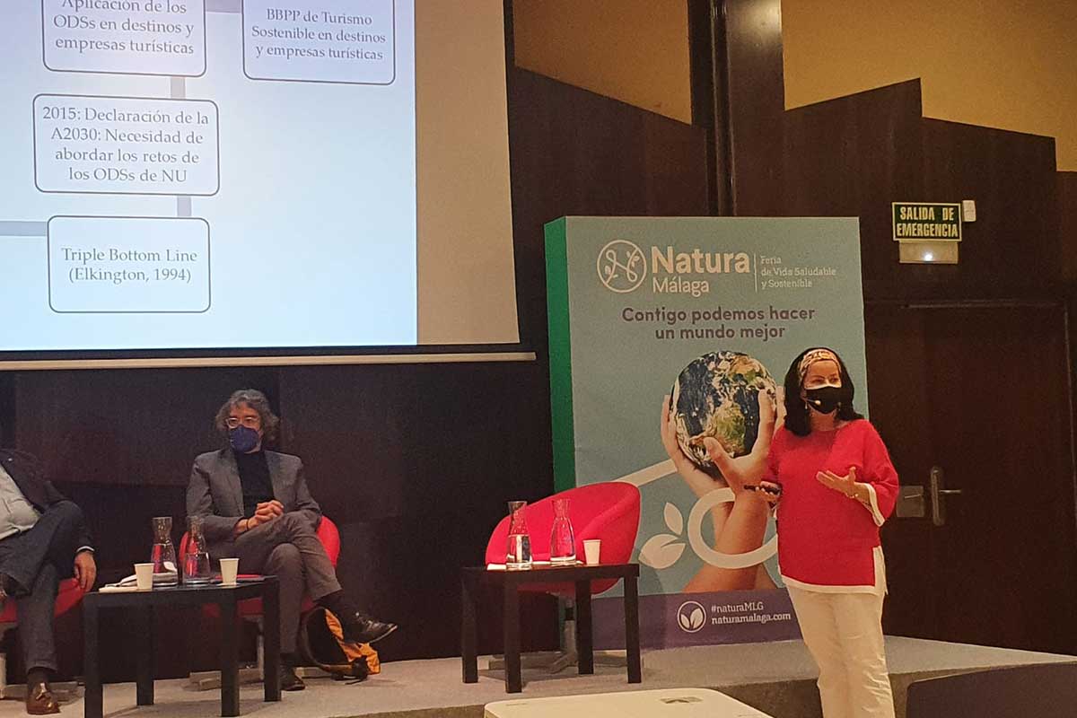 Una mujer con camisa roja y mascarilla habla en una conferencia, mientras dos personas sentadas escuchan; al fondo se ve una diapositiva de presentación y una pancarta de Natura Málaga.