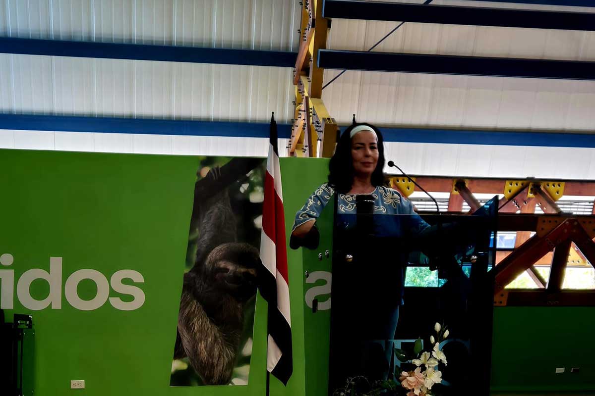 Una mujer está de pie en un podio hablando, con una bandera de Costa Rica y un fondo verde con una imagen de un perezoso y texto parcial visible detrás de ella.