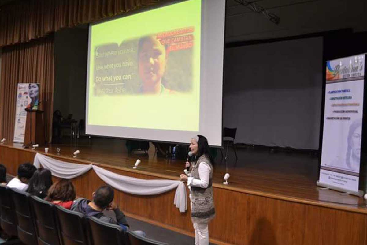 Una mujer habla por un micrófono en el escenario frente a un público sentado, con una presentación proyectada en una gran pantalla detrás de ella.