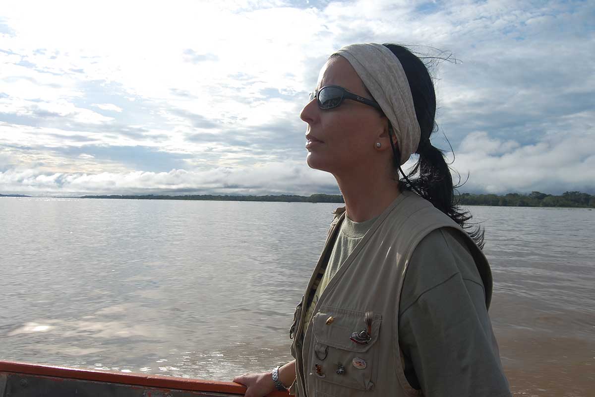 Una mujer con gafas de sol y diadema está de pie en un barco, mirando hacia un ancho río con un cielo nublado de fondo.