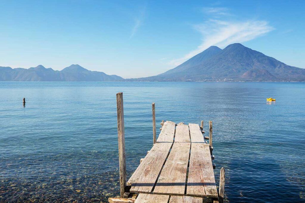 Un muelle de madera se extiende sobre aguas cristalinas hacia montañas lejanas bajo un cielo azul.