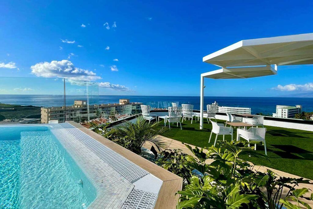 Una terraza en la azotea con piscina, sillas y mesas blancas, vegetación y vistas al océano y a los edificios cercanos bajo un cielo azul despejado.